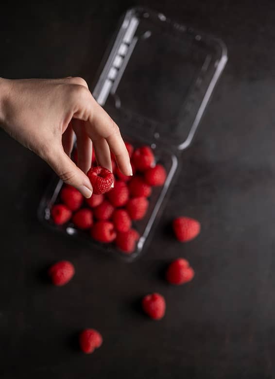 A ladies hand holding a raspberry over a Yucca Packaging punnet clamshell filled with raspberries.