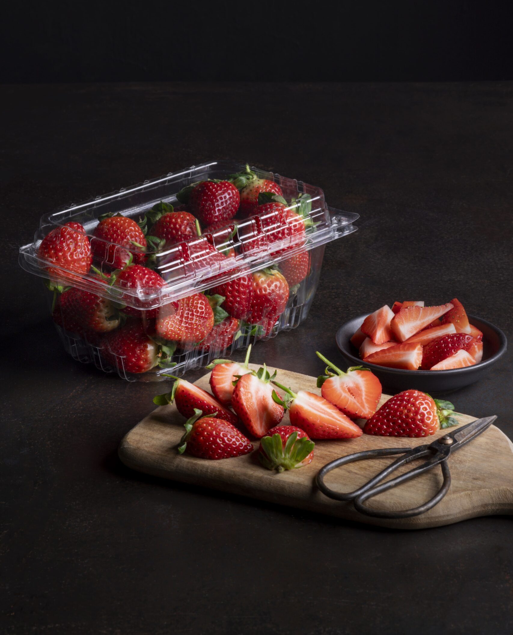 Strawberry punnet with strawberry on a breadboard with a scissor on the side.